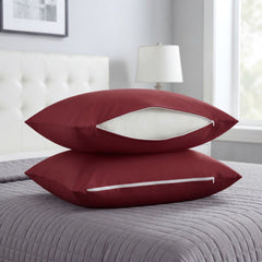 Two burgundy pillowcases stacked on a gray quilted bedspread in a bedroom setting, with a white zipper opening showing a white insert.